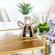The Forged Flare® Burning Candle Ornament is a 3.5" tall Mother's Angels® angel figurine displayed on a gold shelf, with books, a potted succulent, and purple and white flowers in the background.