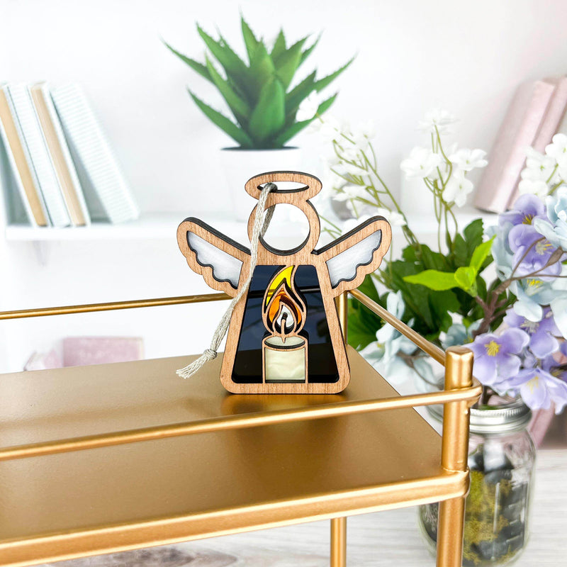 The Forged Flare® Burning Candle Ornament is a 3.5" tall Mother's Angels® angel figurine displayed on a gold shelf, with books, a potted succulent, and purple and white flowers in the background.