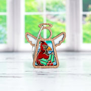 The Forged Flare® Winter Cardinal Ornament with Imperfections is a 3.5" angel figurine from the Mother’s Angels® collection, featuring white wings, a red cardinal centerpiece, and holly detailing, displayed on a white surface before a green garden.