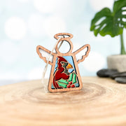 The Forged Flare® Winter Cardinal Ornament with Imperfections, a 3.5" handcrafted Mother's Angels® angel figurine featuring a painted cardinal centerpiece, sits on wood with a green plant and light blue background behind it.
