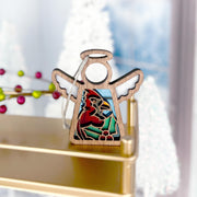 The Winter Cardinal Ornament with Imperfections by Forged Flare® for Mother’s Angels® is a 3.5" wooden angel figurine, featuring a cardinal centerpiece on a gold shelf with frosted trees and red berries in the backdrop.