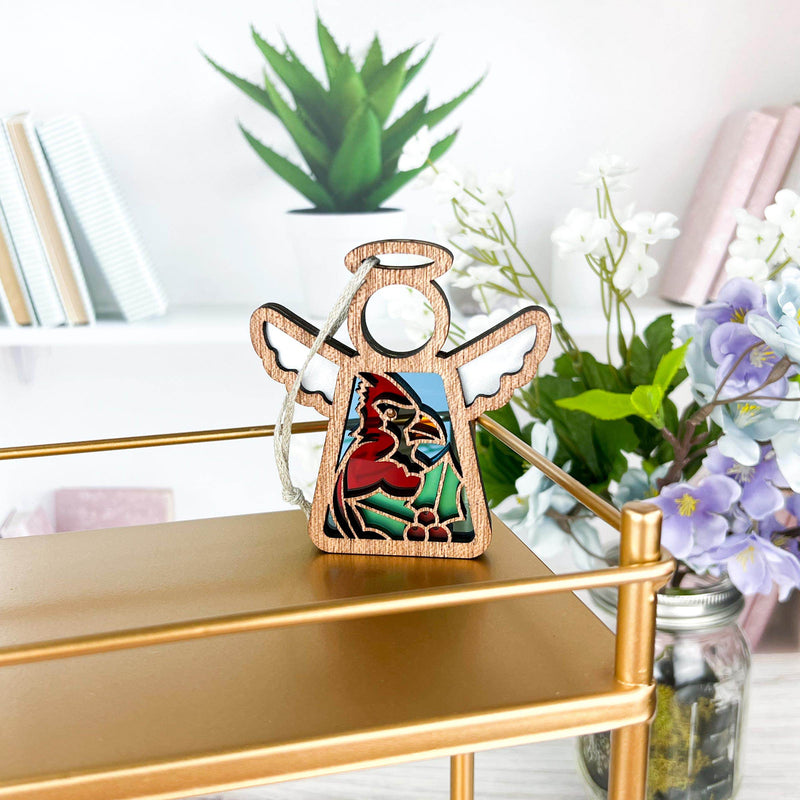 The Forged Flare® Winter Cardinal Ornament with Imperfections—a 3.5" handcrafted Mother's Angels® angel figurine—rests on a gold shelf, backed by potted plants, purple blooms in a jar, and books on white shelves in a bright room.