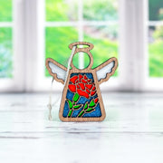 The Carnation Ornament by Forged Flare® is a 3.5" Mother's Angels® figurine featuring stained glass-style wings and a red carnation design, displayed on a white surface with a blurred window and green outdoor background.