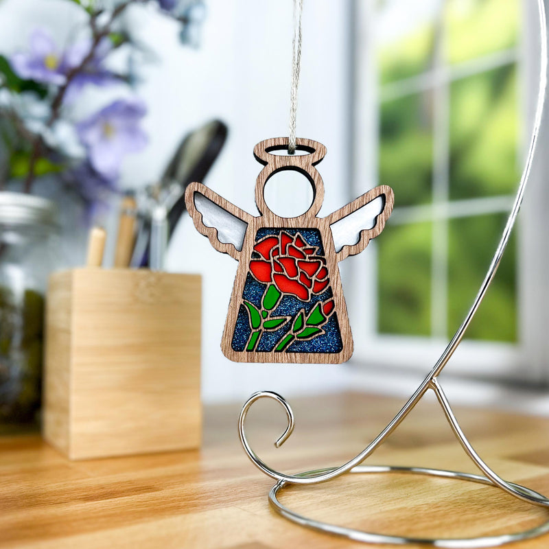 The Forged Flare® Carnation Ornament | 3.5" (H) Angel Figurine from the Mother's Angels® collection hangs from a stand on a wooden desk, with a blurred window and a pen holder in the background.