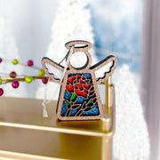 The Forged Flare® Mother's Angels® Carnation Ornament, a 3.5" wooden angel figurine with white wings and a stained glass-style carnation, stands on a gold surface amid blurred holiday decor and twinkling lights.