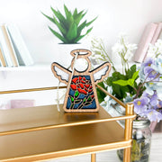 A Forged Flare® Carnation Ornament | 3.5" (H) Mother's Angels® angel figurine with a stained glass-style red rose design sits on a gold shelf, with Carnation flowers, jars, books, and a white wall in the background.