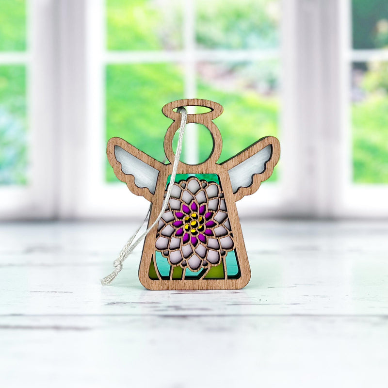 The Forged Flare® Chrysanthemum Ornament with Imperfections is a 3.5" handcrafted Mother's Angels® wooden angel figurine featuring white wings and a colorful chrysanthemum centerpiece, shown on a bright, greenery-filled white surface.