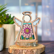 The Forged Flare® Mother's Angels® Chrysanthemum Ornament with Imperfections is a 3.5" handcrafted wooden angel figurine, with white wings and a stained-glass style chrysanthemum centerpiece, displayed on a tree slice beside a potted plant.