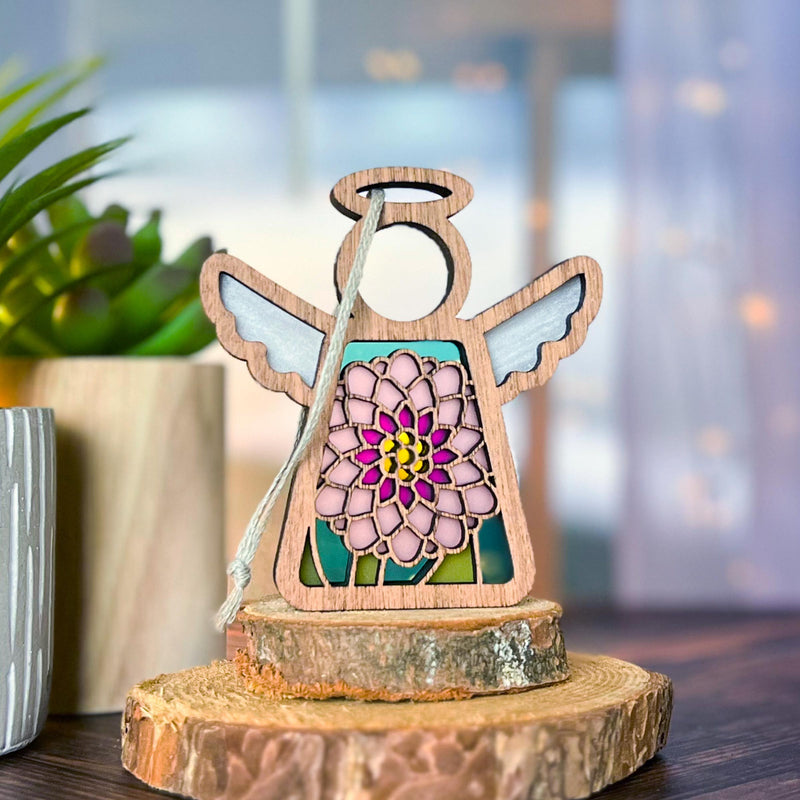 The Forged Flare® Mother's Angels® Chrysanthemum Ornament with Imperfections is a 3.5" handcrafted wooden angel figurine, with white wings and a stained-glass style chrysanthemum centerpiece, displayed on a tree slice beside a potted plant.