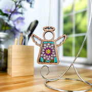 The Chrysanthemum Ornament with Imperfections—a 3.5" handcrafted angel figurine by Forged Flare® Mother's Angels®—features stained glass-style florals and hangs from a metal stand on a wooden table, with flowers and sunlight in the blurred background.