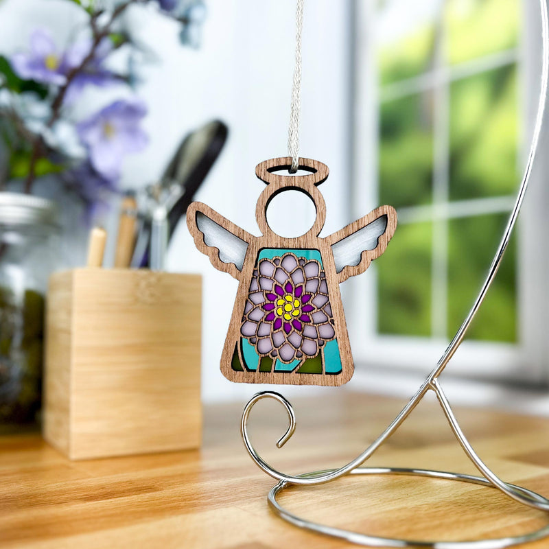 The Chrysanthemum Ornament with Imperfections—a 3.5" handcrafted angel figurine by Forged Flare® Mother's Angels®—features stained glass-style florals and hangs from a metal stand on a wooden table, with flowers and sunlight in the blurred background.