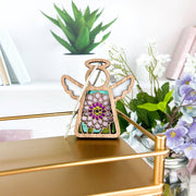 The Forged Flare® Mother's Angels® Chrysanthemum Ornament with Imperfections is a 3.5" handcrafted angel figurine with a colorful flower design, displayed among green plants, purple flowers, and books for a cozy touch.