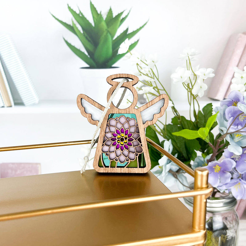 The Forged Flare® Mother's Angels® Chrysanthemum Ornament with Imperfections is a 3.5" handcrafted angel figurine with a colorful flower design, displayed among green plants, purple flowers, and books for a cozy touch.