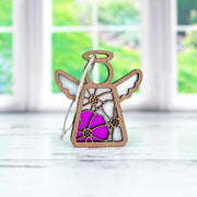 The Forged Flare® Cosmos Ornament | 3.5" (H) Mother's Angels® figurine features a wooden angel with cut-out wings and a pink cosmos flower design, displayed on a light surface with a blurred window and greenery behind it.