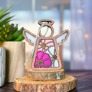 The Forged Flare® Cosmos Ornament, a 3.5" Mother's Angels® wooden angel figurine with stained glass-style flowers, stands on a tree slice beside potted greens, set against a softly blurred background.