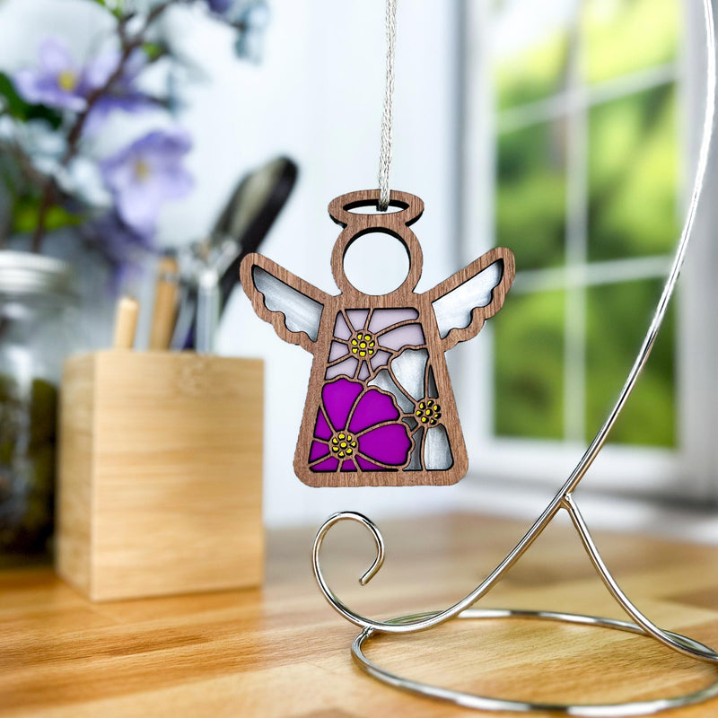 The Cosmos Ornament 3.5" Angel Figurine by Forged Flare® Mother's Angels® stands on a curved metal stand on a wooden desk, featuring stained glass-style wings and a cosmos flower, with flowers and a window blurred in the background.