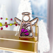 A Cosmos Ornament | 3.5" Angel Figurine from the Mother's Angels® collection by Forged Flare® stands on a gold shelf. This wooden angel ornament showcases a stained glass design featuring purple, pink, and white cosmos flowers. In the background, there are white, snow-covered trees and a blurred wintry scene. The angel has a string for hanging.