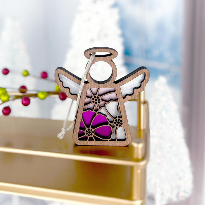 A Cosmos Ornament | 3.5" Angel Figurine from the Mother's Angels® collection by Forged Flare® stands on a gold shelf. This wooden angel ornament showcases a stained glass design featuring purple, pink, and white cosmos flowers. In the background, there are white, snow-covered trees and a blurred wintry scene. The angel has a string for hanging.