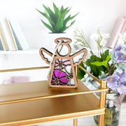 A Forged Flare® Mother's Angels® 3.5" cosmos ornament angel figurine with pink and purple stained glass-style flowers sits on a gold shelf amid books and birth flower decor, with greenery in the background.