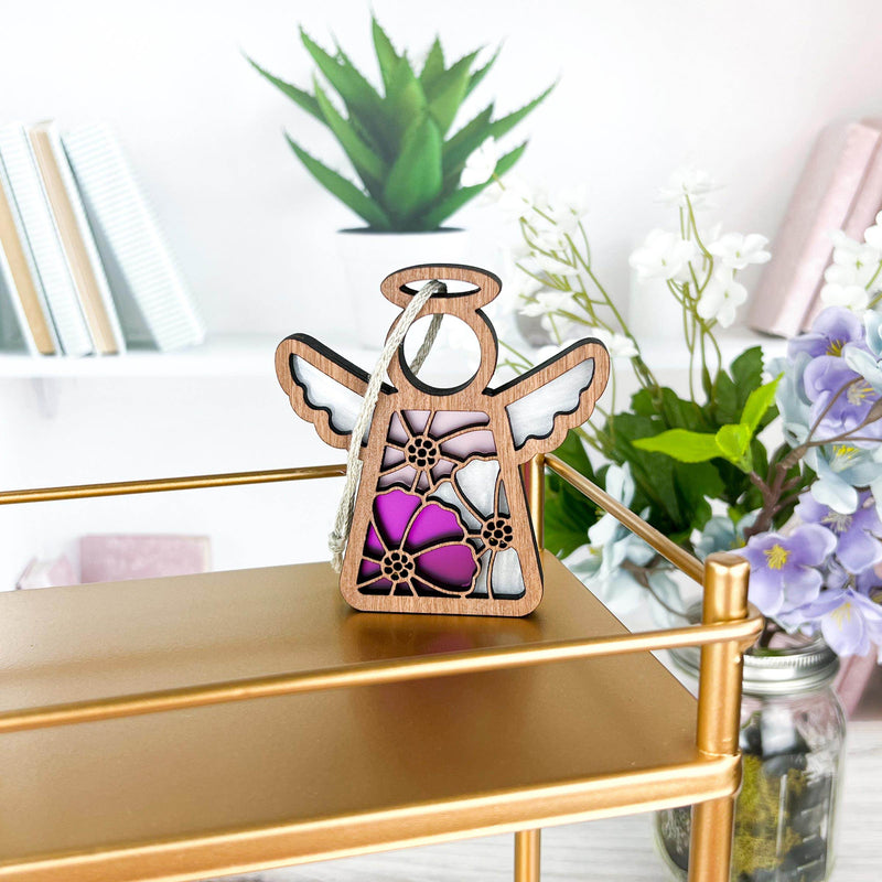 A Forged Flare® Mother's Angels® 3.5" cosmos ornament angel figurine with pink and purple stained glass-style flowers sits on a gold shelf amid books and birth flower decor, with greenery in the background.