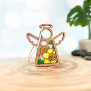 The Forged Flare® Daffodil Ornament | 3.5" (H) Mother's Angels® angel figurine features stained-glass-style daffodils, white wings, and a halo—perfect as a birth flower gift. A blurred green plant is seen in the background.