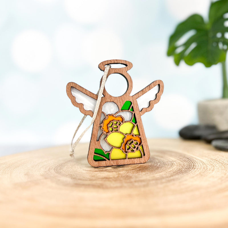 The Forged Flare® Daffodil Ornament | 3.5" (H) Mother's Angels® angel figurine features stained-glass-style daffodils, white wings, and a halo—perfect as a birth flower gift. A blurred green plant is seen in the background.