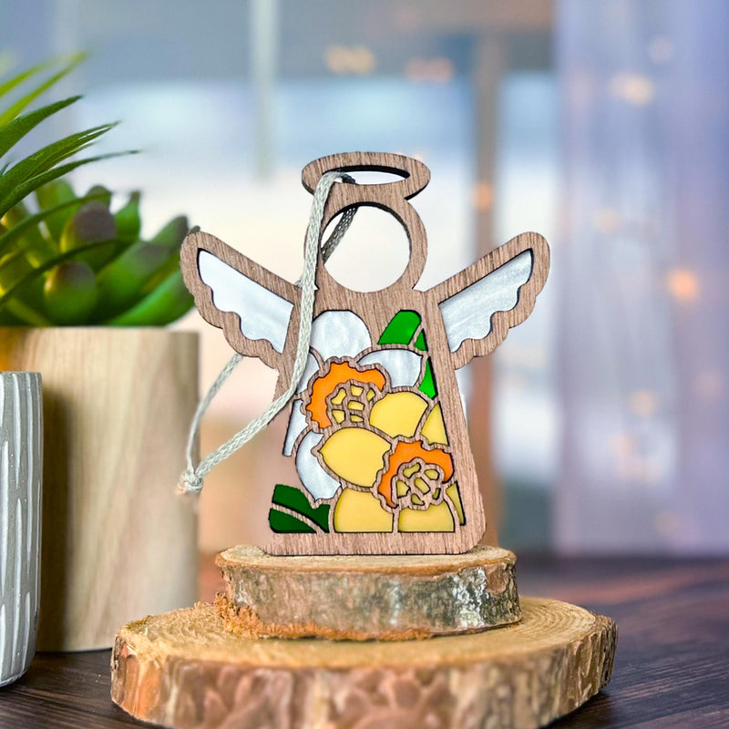 The Daffodil Ornament, a 3.5" angel figurine with white wings and daffodil flowers by Forged Flare® Mother's Angels®, stands on a wooden slice with a potted plant and curtains behind—an ideal birth flower gift.