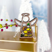 A Forged Flare® Mother's Angels® daffodil ornament, a 3.5" wooden angel figurine with stained glass-style wings, hangs on a gold shelf with blurred white Christmas trees and festive berries in the background.