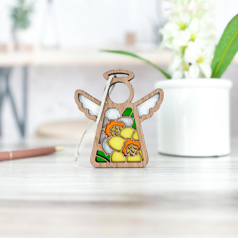 A Forged Flare® Mother's Angels® Daffodil Ornament, a 3.5" angel figurine hand-painted with daffodils, stands on a light table; a blurry white pot and pen in the background make this a thoughtful birth flower gift.