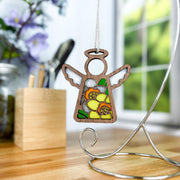 A Forged Flare® Daffodil Ornament | 3.5" (H) Angel Figurine from the Mother's Angels® collection hangs from a silver stand on a desk, with blurred flowers and a window in the background—an ideal birth flower gift.