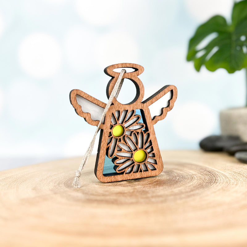 A Forged Flare® Warehouse Return - Daisy Ornament | 3.5" (H) Mother's Angels® figurine with cut-out wings and daisies stands on a smooth surface, a silver string atop, with a green leaf and stones softly blurred behind.