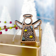 A Warehouse Return - Daisy Ornament (3.5" H) Mother's Angels® angel figurine by Forged Flare®, with white wings and yellow daisies, stands on a gold surface, with blurred white Christmas trees and red berries in the background.
