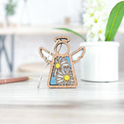 A Warehouse Return - Daisy Ornament | 3.5" (H) Angel Figurine by Forged Flare® from the Mother's Angels® line sits on a wooden surface, with a blurred pen and plant in the background—ideal for gifting or selling on Amazon.