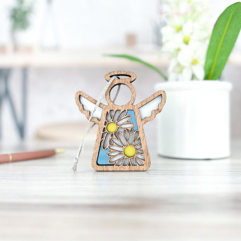 A Warehouse Return - Daisy Ornament | 3.5" (H) Angel Figurine by Forged Flare® from the Mother's Angels® line sits on a wooden surface, with a blurred pen and plant in the background—ideal for gifting or selling on Amazon.