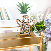 A 3.5" Mother's Angels® Daisy Ornament by Forged Flare® sits on a gold shelf with books, a green potted plant, and white and purple flowers in the background.
