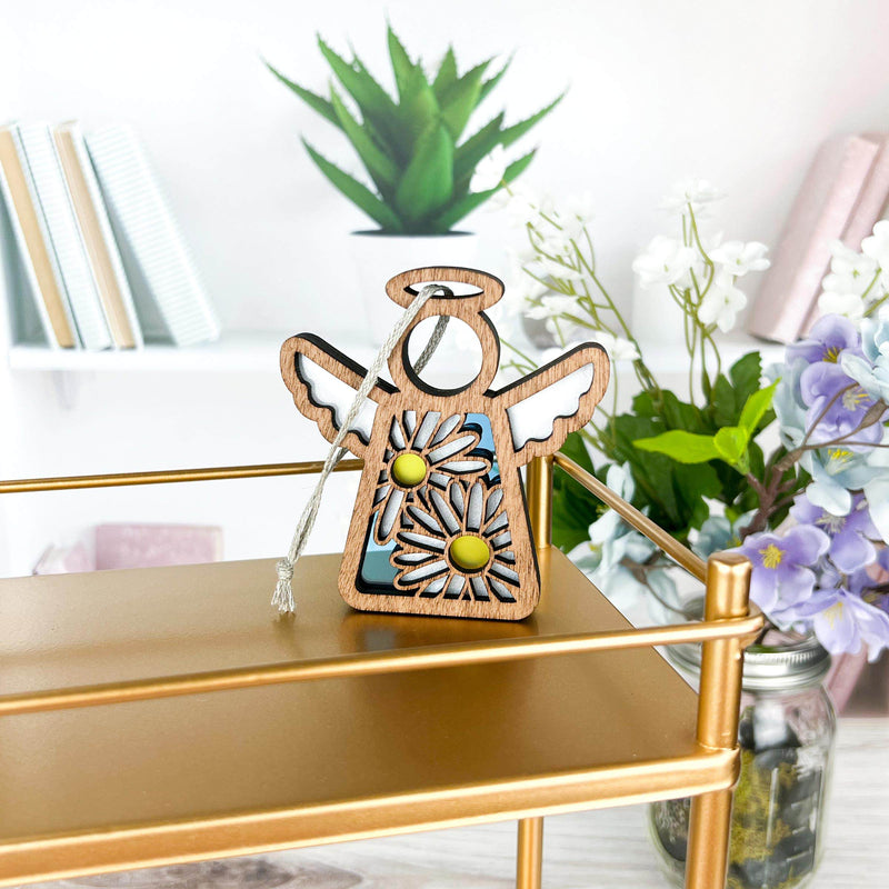 A 3.5" Mother's Angels® Daisy Ornament by Forged Flare® sits on a gold shelf with books, a green potted plant, and white and purple flowers in the background.