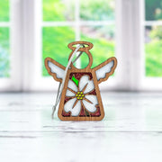 The Forged Flare® Dogwood Ornament is a 3.5" Mother's Angels® figurine featuring white wings and a floral dogwood design, displayed on a white surface with a blurred window and greenery in the background.