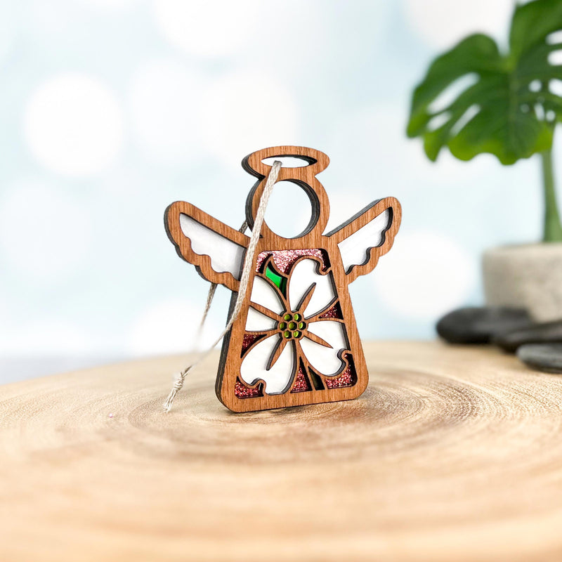 The Forged Flare® Dogwood Ornament is a 3.5" wooden Mother's Angels® figurine with white wings and a dogwood flower, perfect as a Christmas tree ornament. It stands on wood, with a blurred plant and stacked stones in the background.
