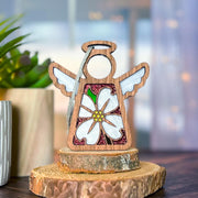 The Forged Flare® Mother's Angels® Dogwood Ornament is a 3.5" wooden angel figurine with white wings and a dogwood flower, displayed on a small tree stump against a soft, green background.
