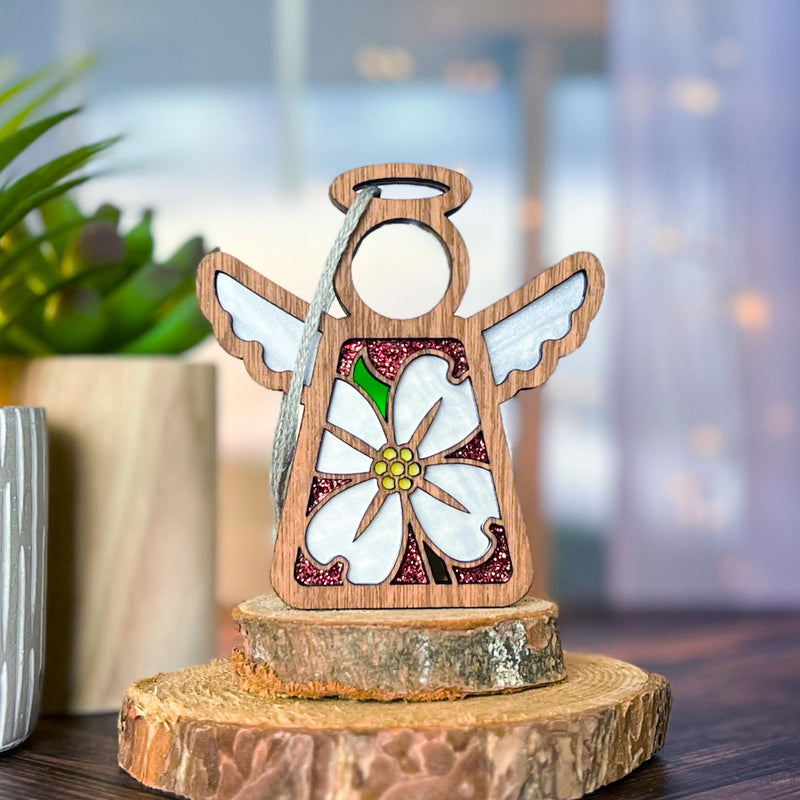 The Forged Flare® Mother's Angels® Dogwood Ornament is a 3.5" wooden angel figurine with white wings and a dogwood flower, displayed on a small tree stump against a soft, green background.