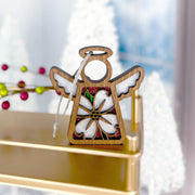 The Forged Flare® Dogwood Ornament | 3.5" (H) Mother's Angels® figurine features a wooden angel with white wings and a dogwood flower, accented in red glitter, sitting on a gold shelf with blurred holiday decorations and white Christmas trees behind.