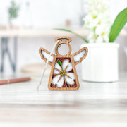 The Dogwood Ornament by Forged Flare®, part of the Mother's Angels® collection, is a beautiful 3.5" wooden angel figurine with wings outspread, showcasing an elegant flower design in the center. This Christmas tree ornament hangs from a delicate white string. In the softly focused background, green leaves, white dogwood flowers, and a pen resting on a surface can be seen.