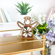 The Forged Flare® Dogwood Ornament, a 3.5" Mother's Angels® angel figurine, hangs by a string on a gold shelf surrounded by pastel books and artificial flowers.