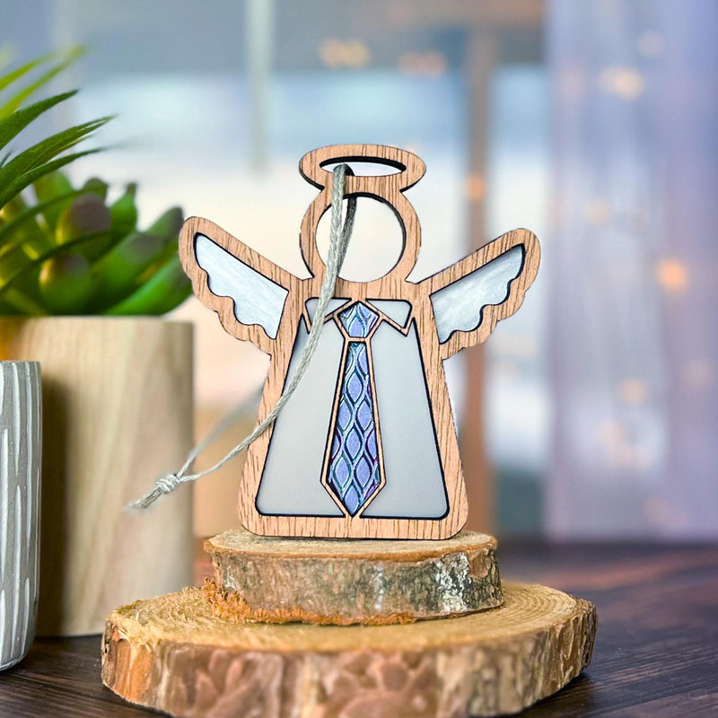 The Forged Flare® Forever Your Father Ornament, a 3.5" Mother's Angels® wooden angel figurine with a blue tie and white wings, is displayed on a tree slice near a small plant—ideal as a Christmas ornament or Father's Day gift. Blurred lights and window in background.