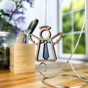 The Forged Flare® Forever Your Father Ornament | 3.5" Mother's Angels® figurine with a blue tie hangs on a metal stand—ideal for Father's Day. Set on a wooden desk with pens, box, flowers, and greenery outside the window.