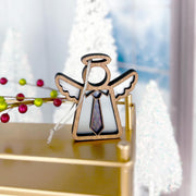 The Forged Flare® Forever Your Father Ornament (3.5" H) by Mother's Angels® features a wooden angel with halo, wings, and necktie on a gold surface—perfect for Father’s Day or Christmas. Blurred ornaments and white trees appear in the background.