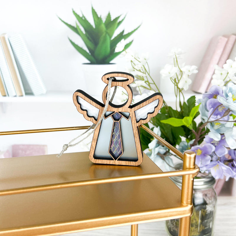 The Forever Your Father Ornament by Forged Flare® is a 3.5" wooden angel figurine with a necktie, hanging by a string on a gold shelf amid books, flowers, and a plant—ideal for Christmas or Father's Day by Mother's Angels®.