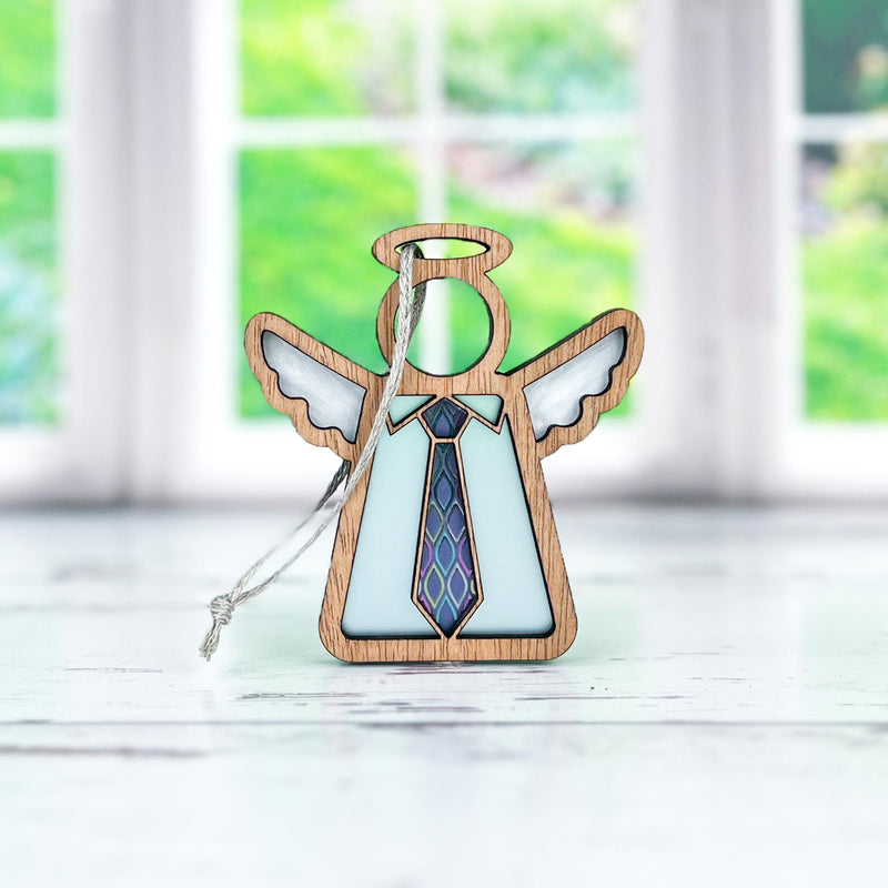 The Forged Flare® Forever Your Father Ornament, a 3.5" Mother's Angels® figurine with white wings and a blue patterned tie, stands on a white surface—perfect for Father's Day—with a blurred window and green background behind.