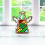 The Forged Flare® Mother's Angels® 3.5" wooden angel figurine features a rainbow stained glass body and a glittery four-leaf clover ornament at its heart, displayed on a white surface by a bright window.