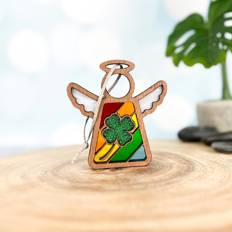 The Glittery Four-Leaf Clover Ornament by Forged Flare® is a 3.5" Mother's Angels® figurine featuring rainbow stripes, white wings, and a sparkling green clover, displayed on wood with a plant and soft-focus background.